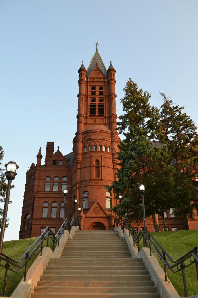A stunning red sandstone Romanesque building, looms over a long concrete staircase. The architectural detail of the bell tower and arched windows is highlighted by a clear, late-afternoon sky.