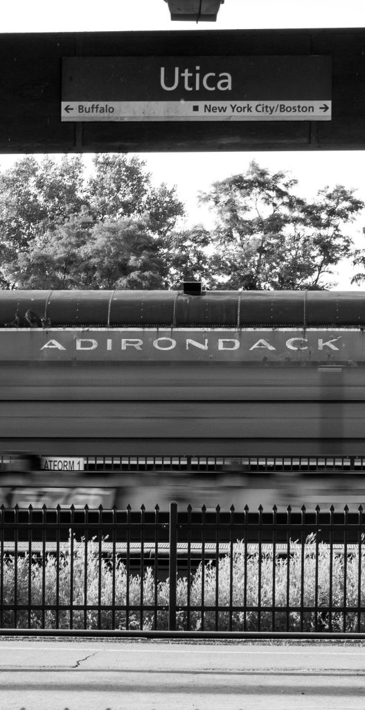 This black-and-white photo captures a train car labeled "ADIRONDACK" at the Utica station. A sign above points toward Buffalo, NYC, and Boston, while motion blur on the lower tracks adds a sense of transit and history to the scene.
