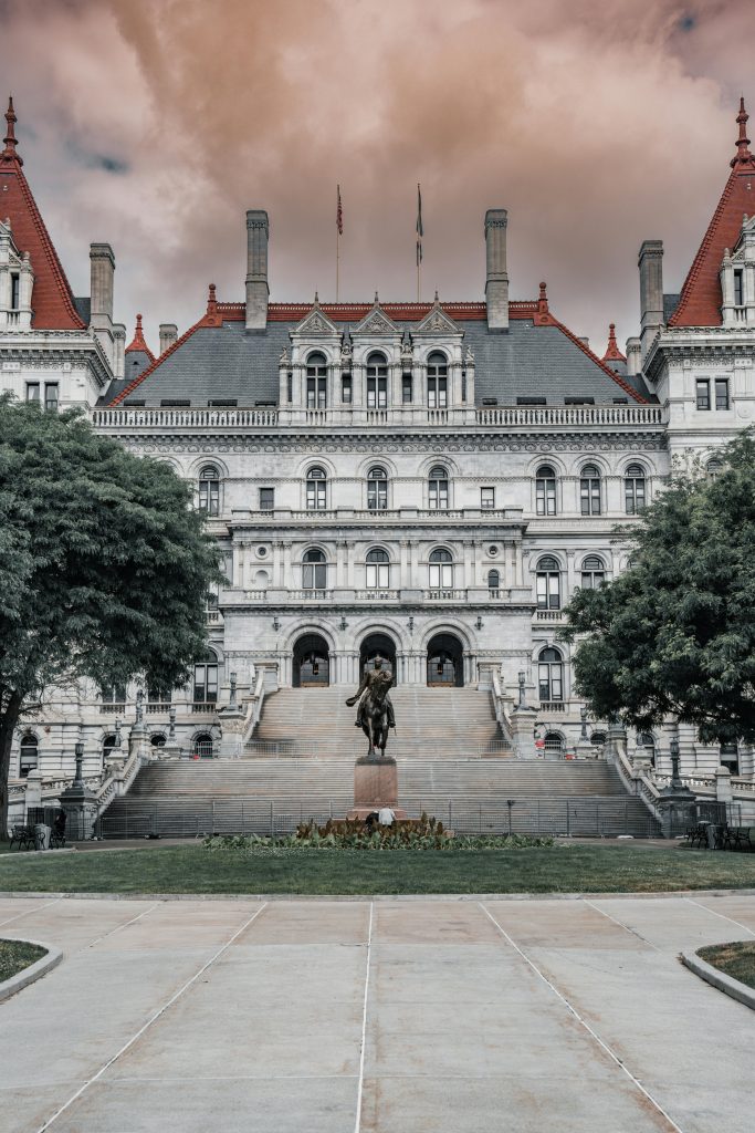 The New York State Capitol in Albany stands grandly under a moody, copper-toned sky. An equestrian statue of Philip Sheridan anchors the wide stone staircase, while the building's intricate French Renaissance architecture creates a regal backdrop.