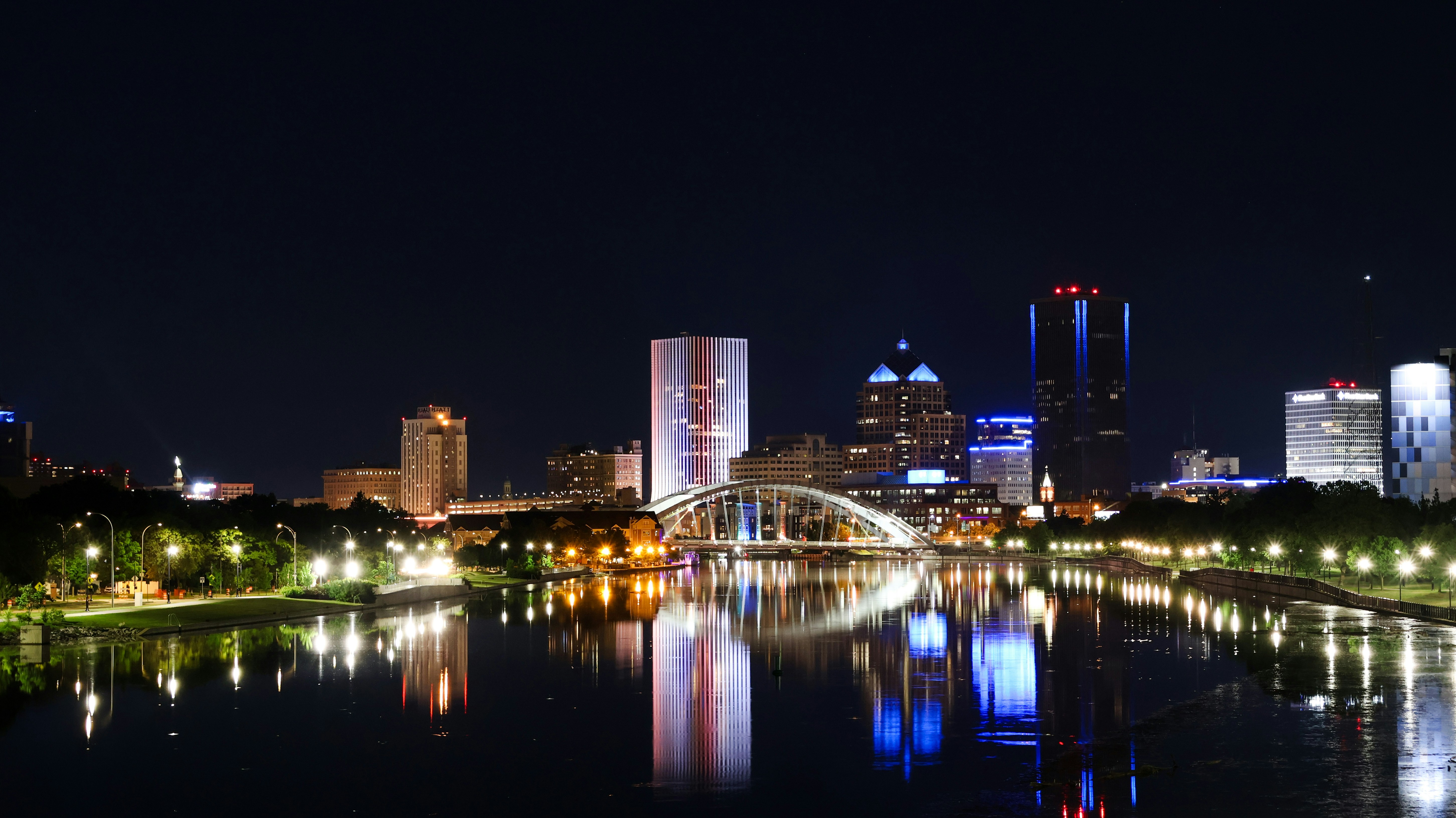 A vibrant nighttime skyline of Rochester, NY, reflects off the Genesee River. The illuminated Frederick Douglass-Susan B. Anthony Memorial Bridge takes center stage, flanked by glowing skyscrapers like the Five Star Bank Plaza and Xerox Tower.